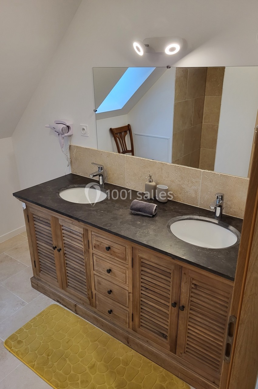 Meuble de salle de bain en bois avec double vasque, miroir, éclairage, et tapis jaune sur un sol carrelé.