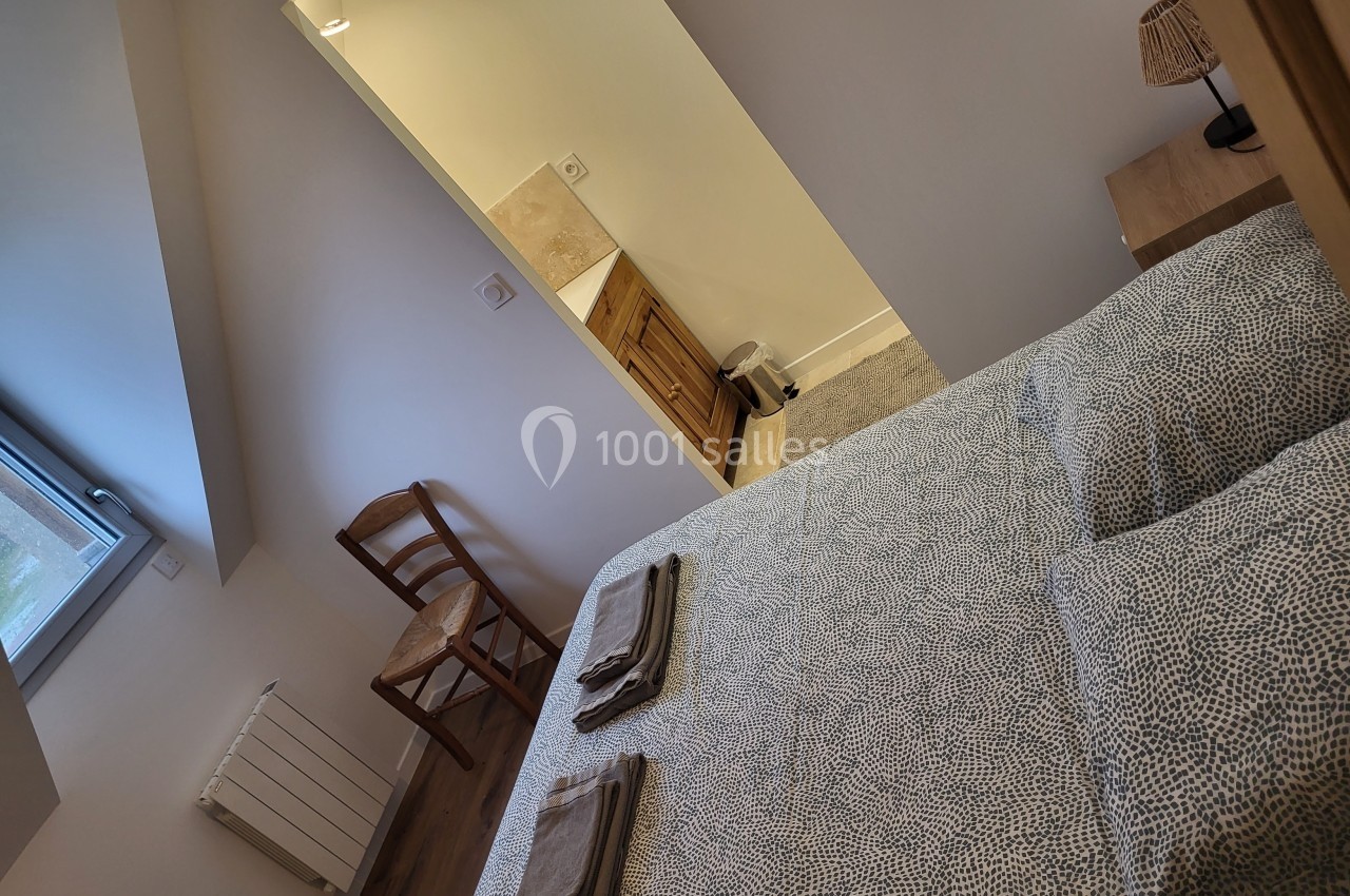 Chambre avec lit double, chaise en bois, fenêtre, et vue sur une salle de bain attenante avec mobilier en bois.