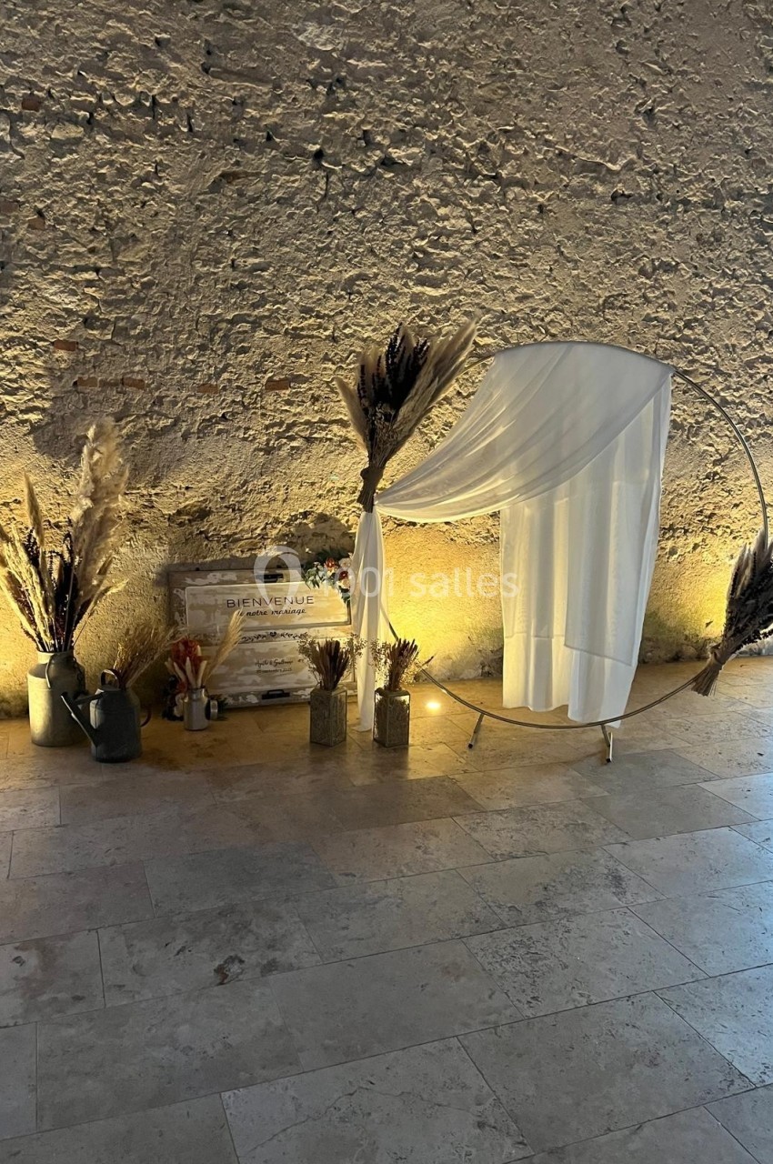 Décoration avec arche circulaire ornée de voilages blancs, pampas et objets rustiques devant un mur en pierre.