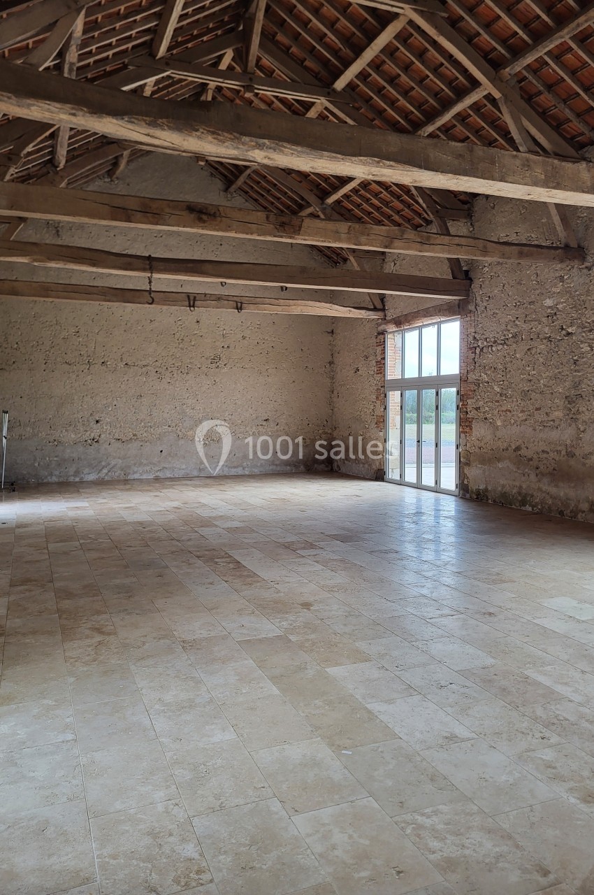Salle vide avec sol en pierre, murs en pierre apparente, poutres en bois au plafond et grande baie vitrée.