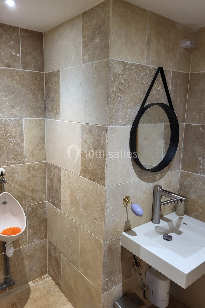 Salle de bain avec lavabo, miroir rond suspendu, urinoir et murs en carrelage beige.