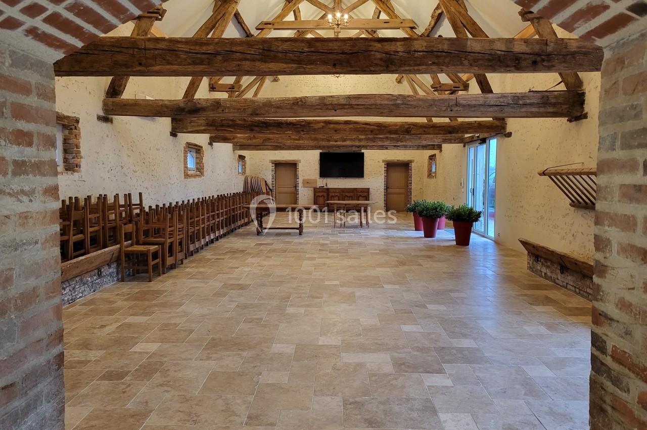 Salle spacieuse avec poutres apparentes, murs en pierre, chaises alignées, tables en bois et plantes en pots rouges.