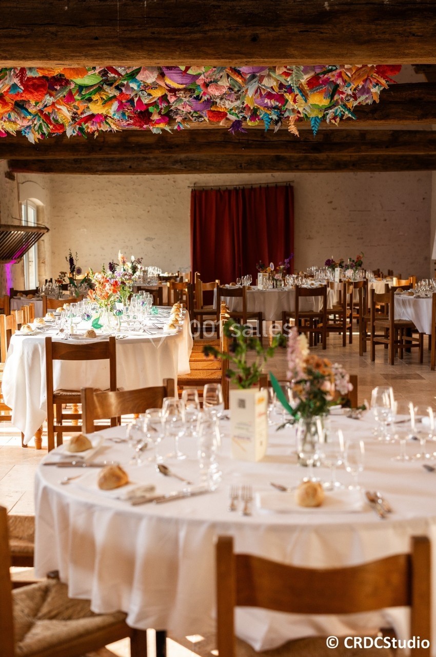Salle de réception lumineuse avec tables rondes dressées, décor coloré suspendu et rideau rouge en arrière-plan.