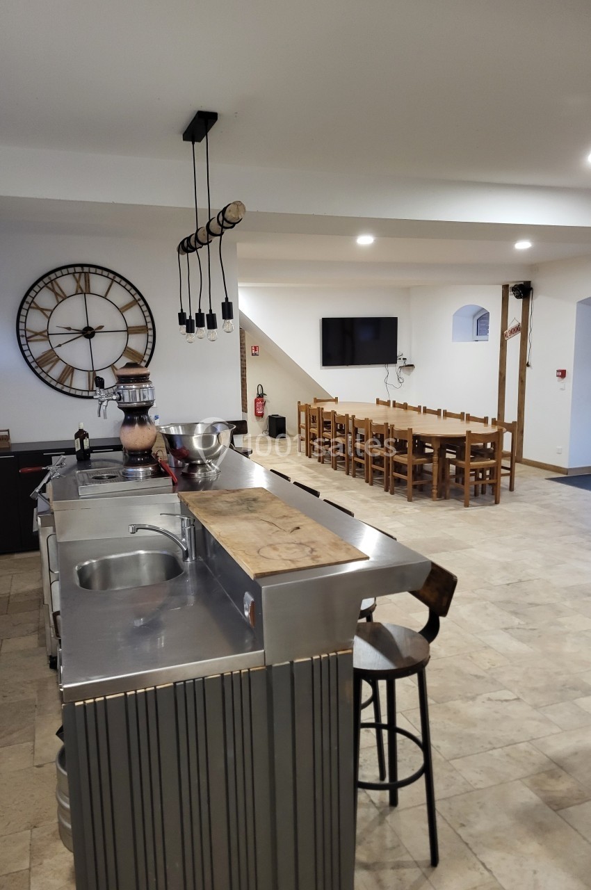 Cuisine ouverte avec comptoir en inox, tabouret, grande horloge murale et salle à manger avec tables en bois.