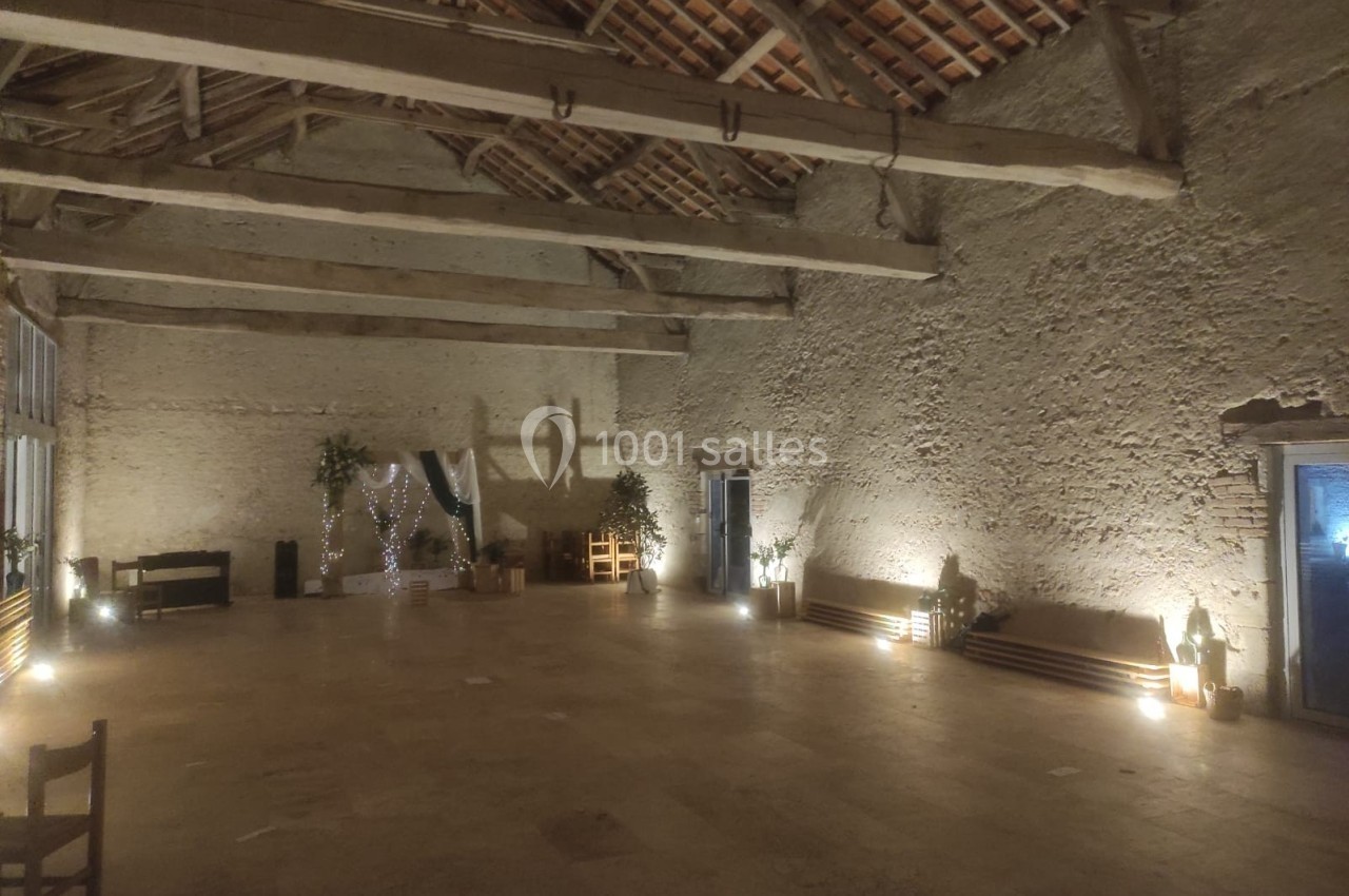 Grande salle en pierre avec poutres apparentes, éclairage tamisé, décorations végétales et guirlandes lumineuses.