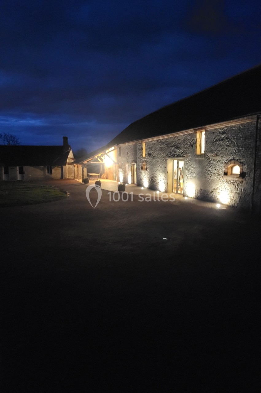 Bâtiment en pierre éclairé par des lumières extérieures, dans une cour sombre au crépuscule.