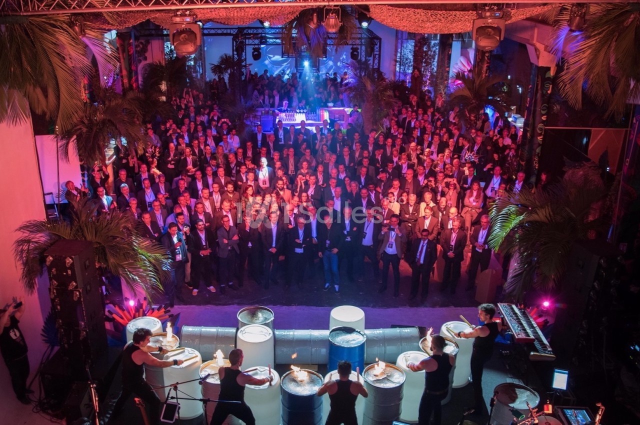 Groupe de musiciens jouant sur des tambours devant une grande foule dans un espace décoré de palmiers et éclairé de lumières…