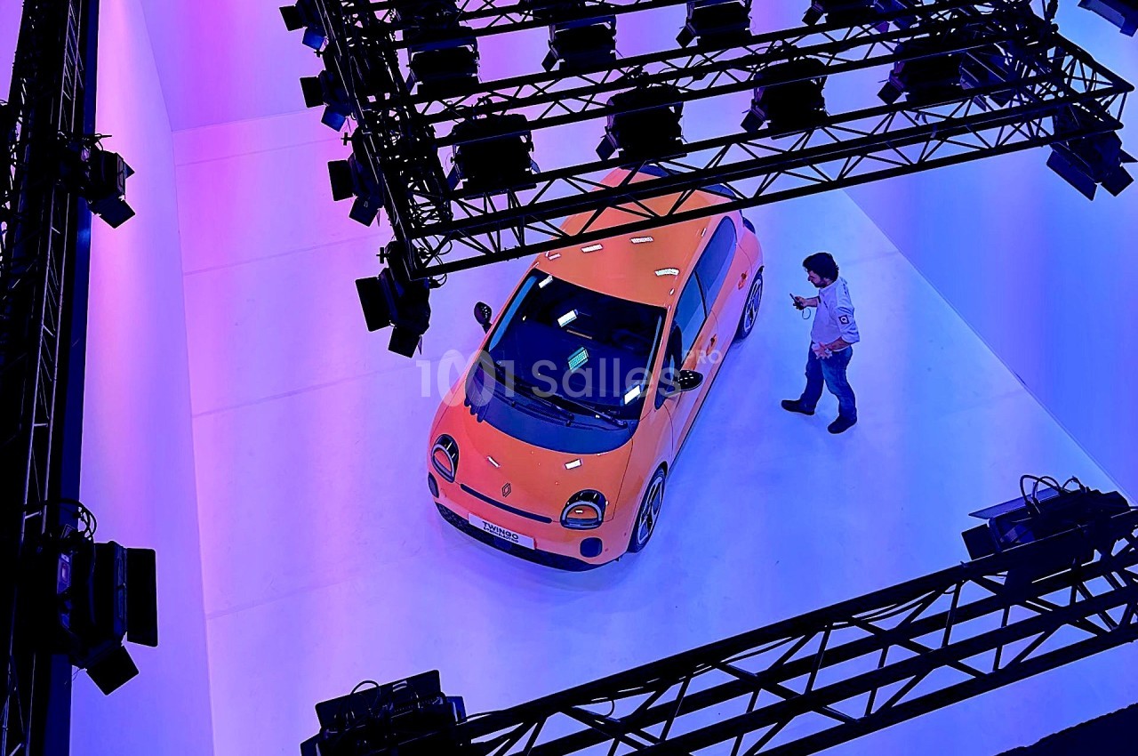 Une voiture orange exposée dans un studio éclairé par des projecteurs violets, avec une personne à proximité.