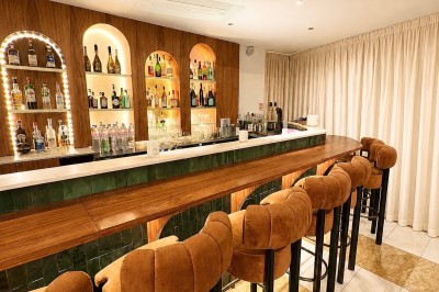 Bar avec comptoir en marbre, tabourets en velours orange et étagères éclairées exposant des bouteilles d'alcool.