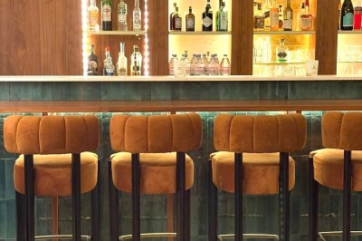 Bar avec comptoir en marbre, tabourets en velours orange et étagères éclairées exposant des bouteilles d'alcool.