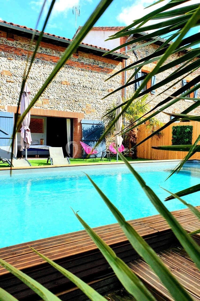 Piscine extérieure entourée de végétation, avec une maison en pierre et des transats en arrière-plan.