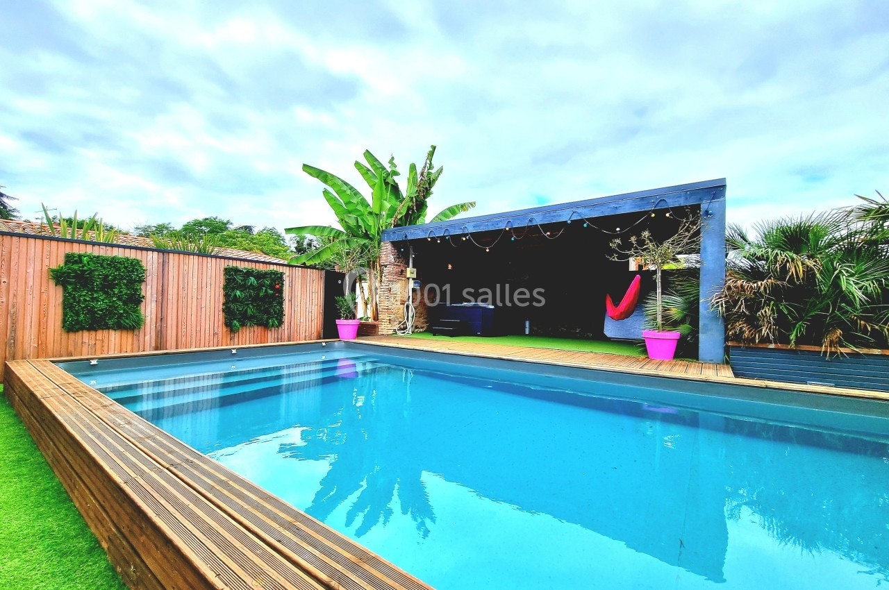 Piscine extérieure entourée de végétation, avec terrasse en bois, hamac et pergola décorée de guirlandes lumineuses.