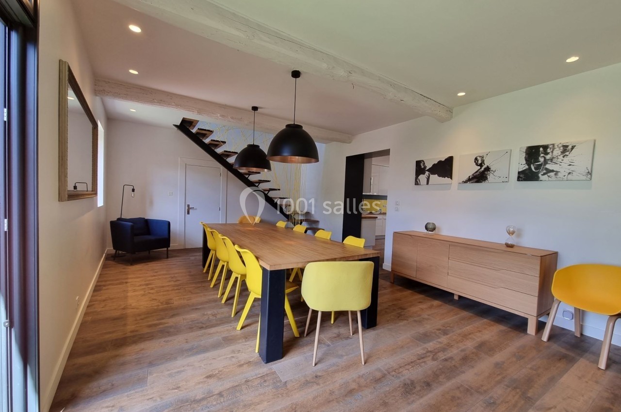Salle à manger moderne avec table en bois, chaises jaunes, suspensions noires et escalier en arrière-plan.