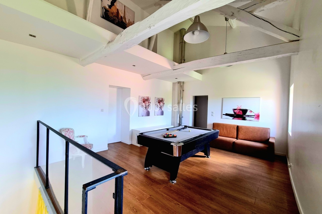 Salle lumineuse avec un billard noir, canapé, fauteuil, poutres apparentes et tableaux décoratifs sur les murs.