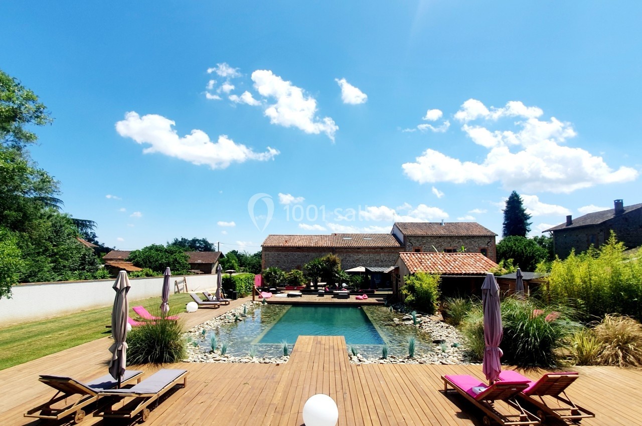 Piscine extérieure entourée de végétation, avec transats et parasols sur une terrasse en bois sous un ciel bleu.