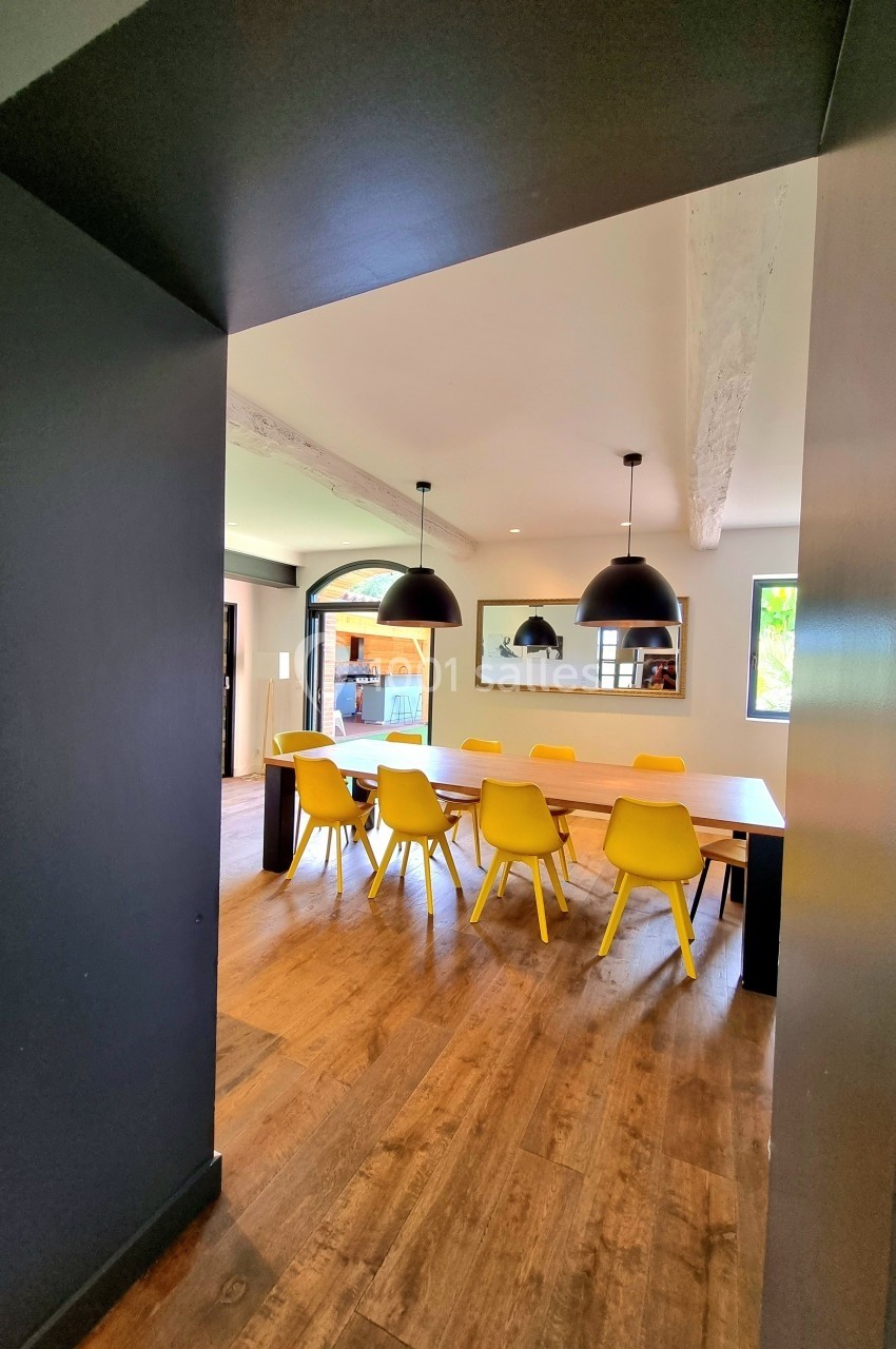 Salle à manger lumineuse avec une table en bois, des chaises jaunes et des suspensions noires, vue sur l'extérieur.