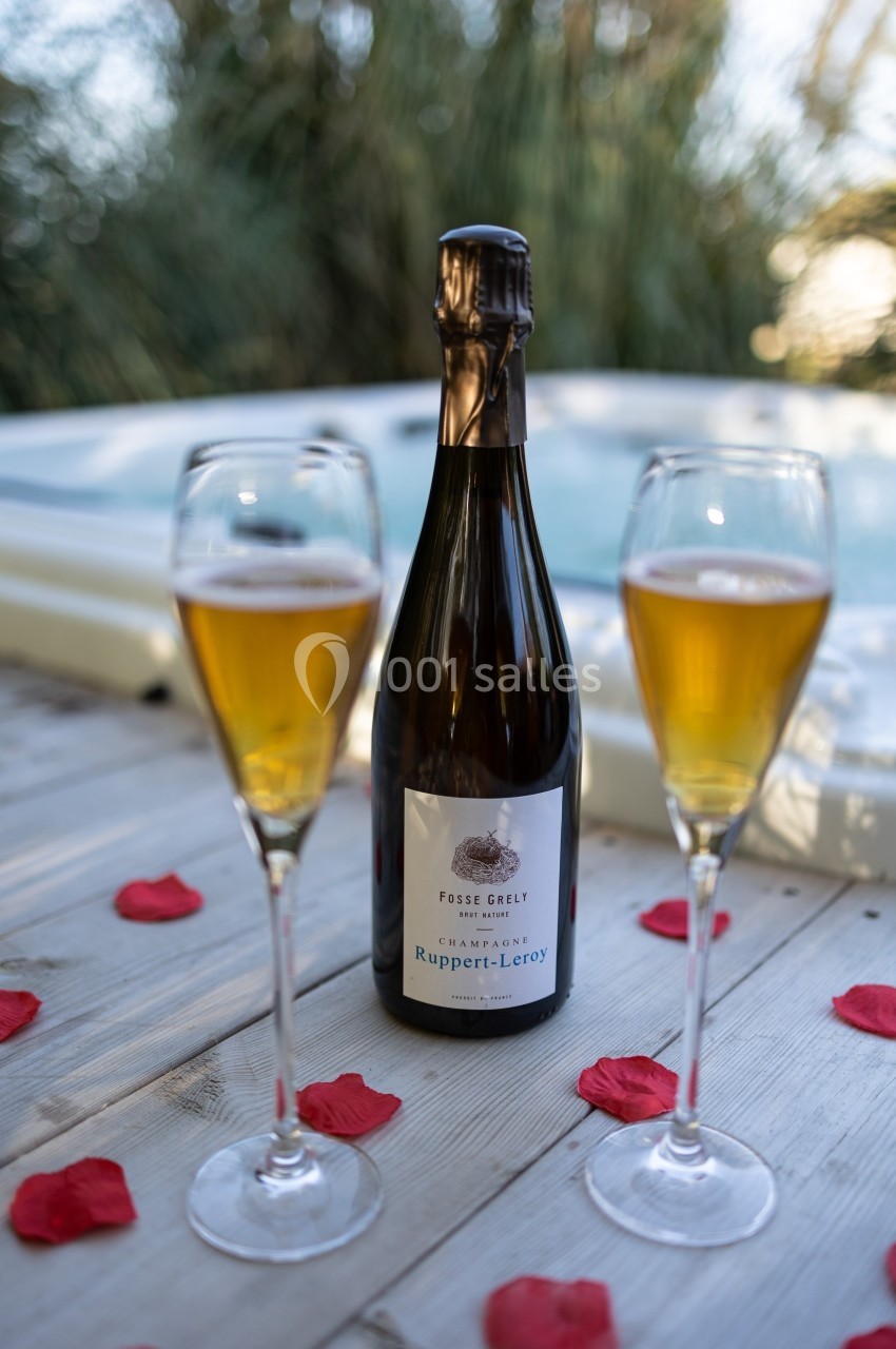 Bouteille de champagne entourée de deux verres remplis, posée sur une table en bois près d'un jacuzzi.