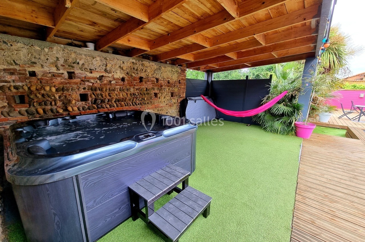 Jacuzzi noir sous un abri en bois, avec hamac rose et sol en gazon synthétique.