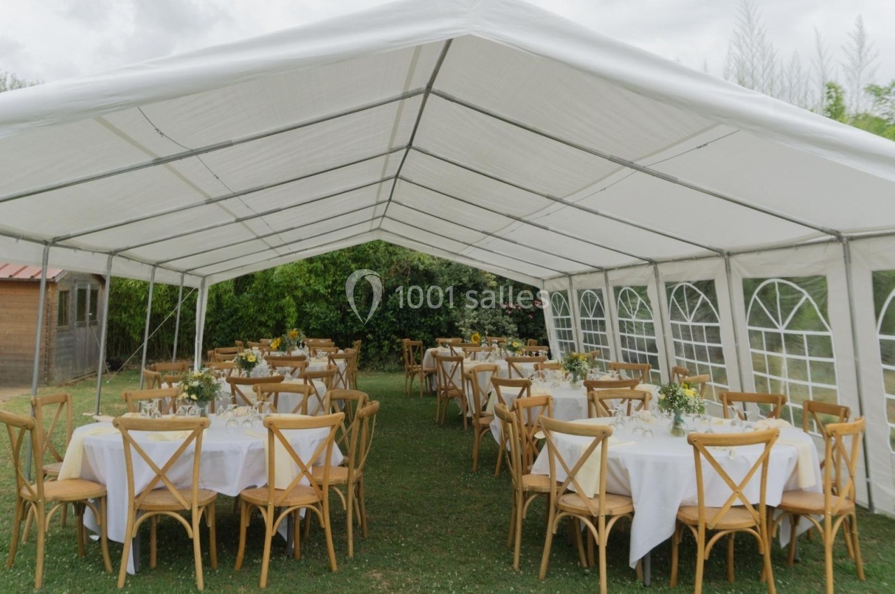 Tente blanche installée en extérieur, abritant des tables rondes dressées avec nappes blanches et chaises en bois.