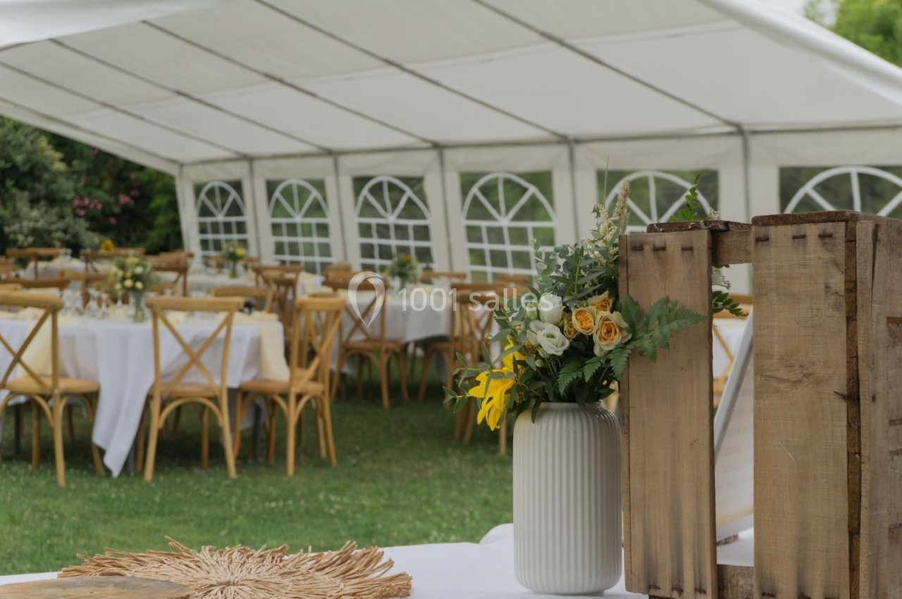 Salle de réception extérieure sous une tente blanche, avec tables dressées et un vase de fleurs au premier plan.