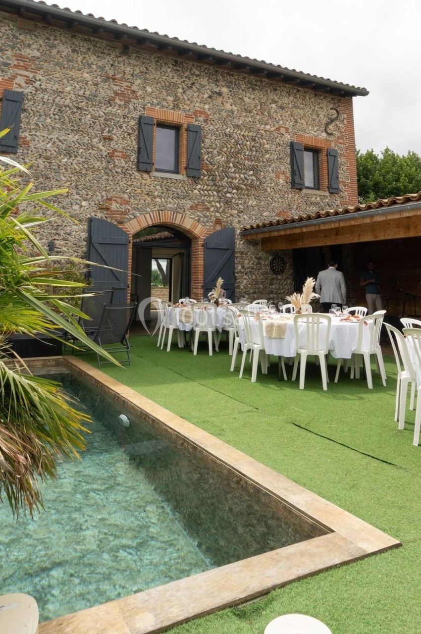 Maison en pierre avec volets bleus, terrasse aménagée pour un repas et piscine bordée de gazon synthétique.