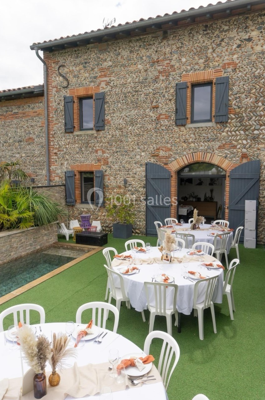 Tables rondes dressées pour un repas en extérieur devant une maison en pierre avec volets bleus et jardin aménagé.