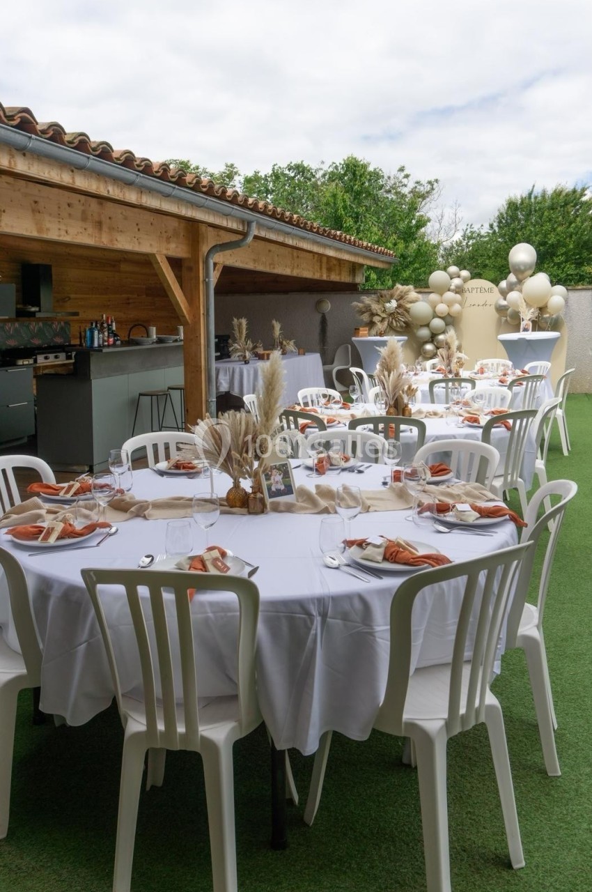 Tables rondes dressées avec nappes blanches, décorations naturelles et chaises blanches, dans un espace extérieur couvert.
