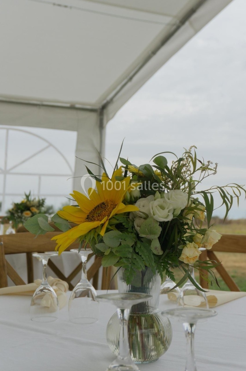 Centre de table avec un bouquet de tournesols, roses blanches et feuillage, posé sur une table dressée sous une tente.