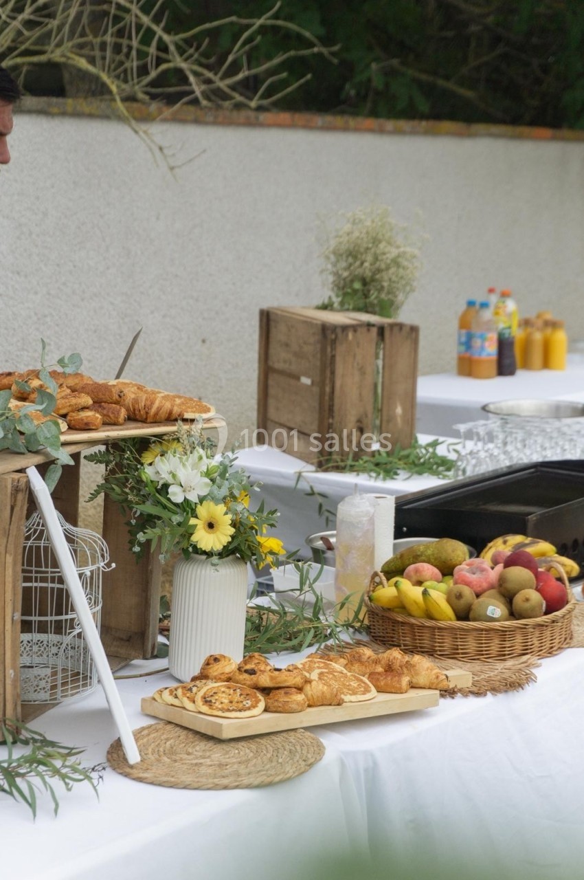 Table de buffet en extérieur avec viennoiseries, fruits, jus de fruits et décoration florale.