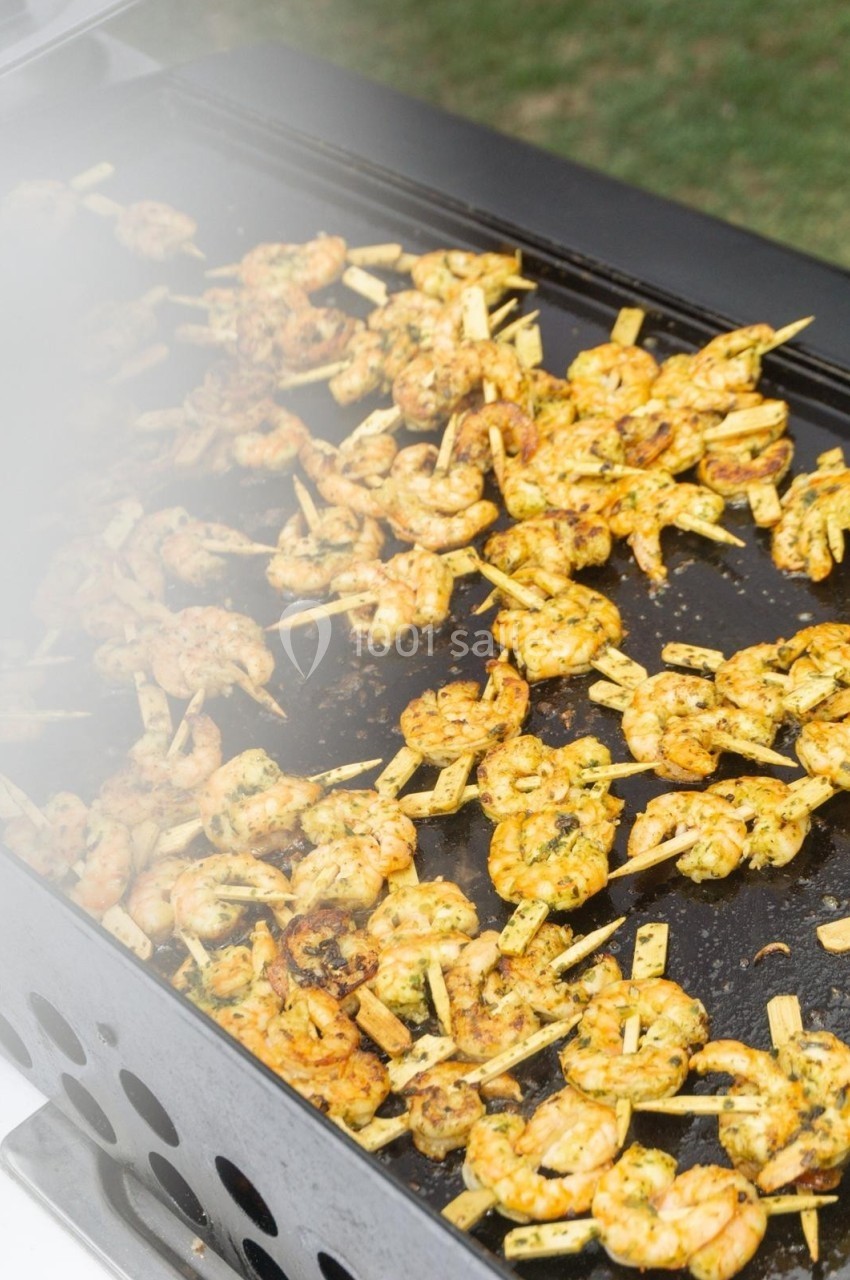 Crevettes marinées grillées sur une plancha, disposées sur des brochettes en bois, en cours de cuisson.