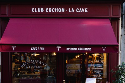 Façade d'un restaurant avec terrasse, auvent rouge et inscriptions ’Club Cochon - La Cave’ et ’Épicerie Cochonne’.
