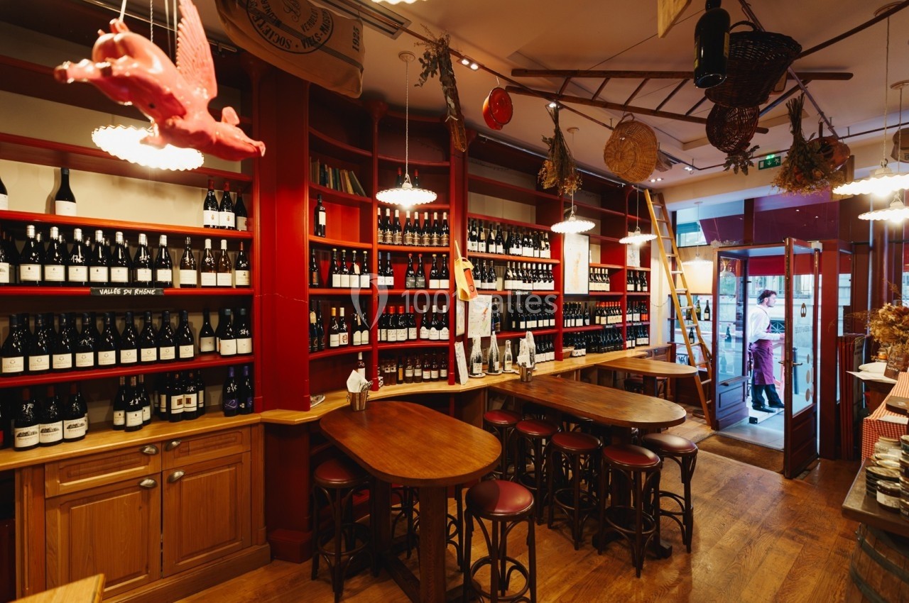 Intérieur d'une cave à vin avec des étagères remplies de bouteilles, tables en bois et décoration suspendue.
