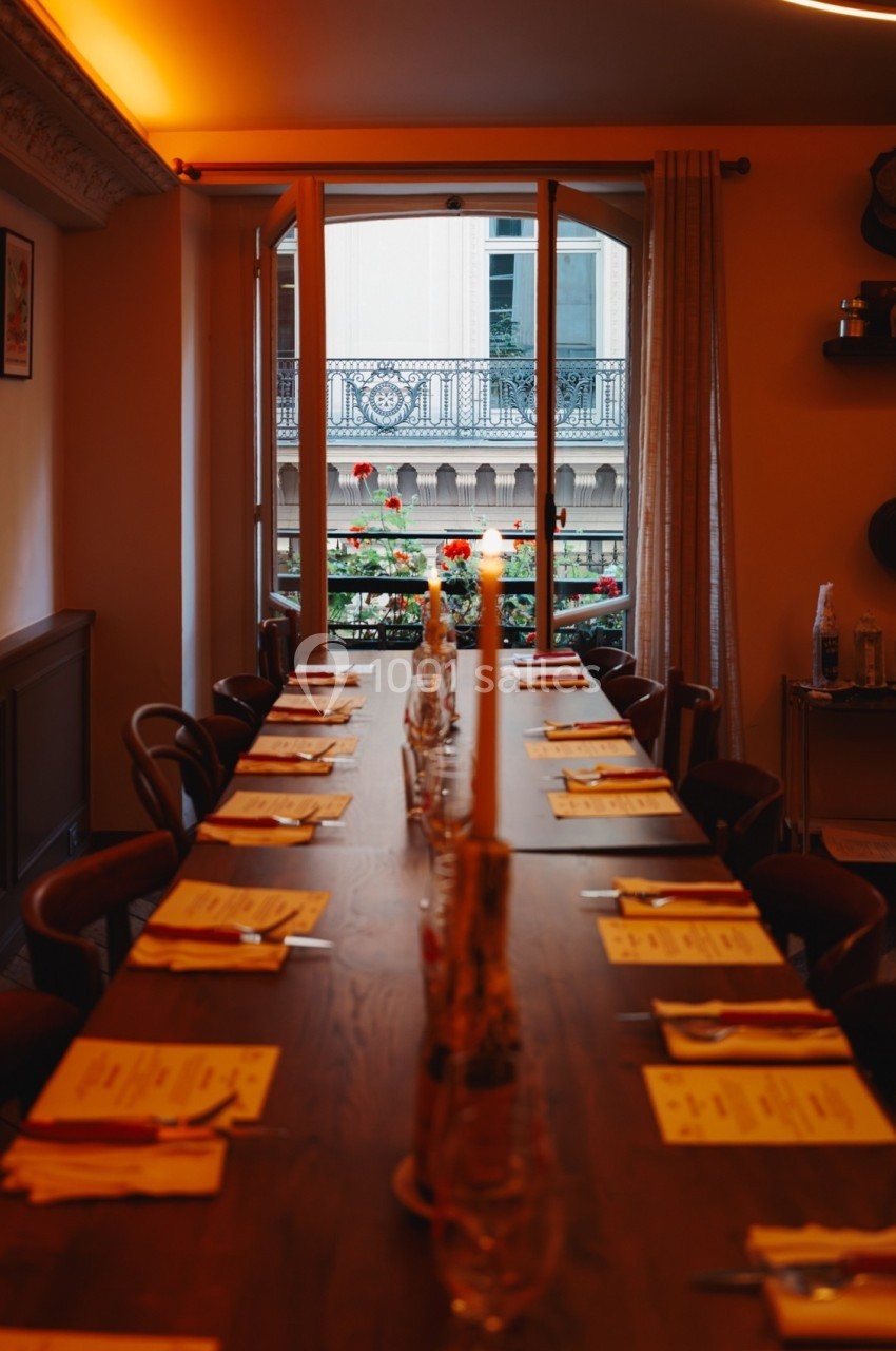 Table en bois dressée avec des menus et des bougies, située dans une salle lumineuse avec vue sur un balcon fleuri.