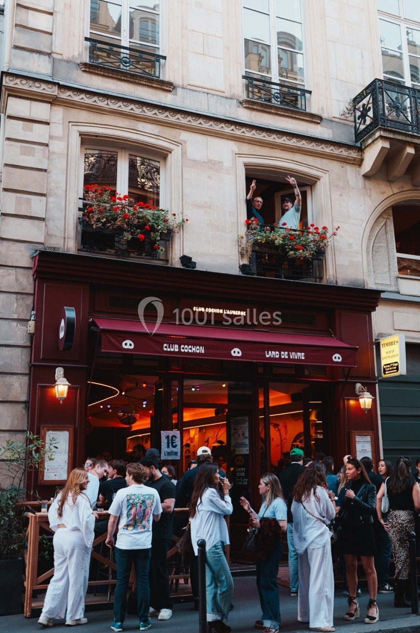 Façade d'un bar animé avec des clients rassemblés en terrasse et deux personnes saluant depuis une fenêtre au-dessus.