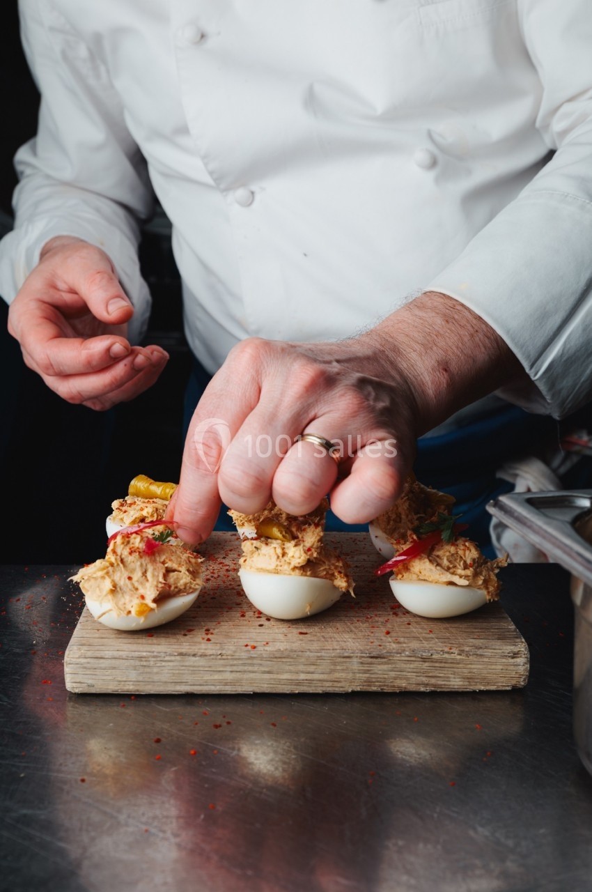 Un chef garnit des œufs farcis avec des condiments sur une planche en bois dans une cuisine professionnelle.