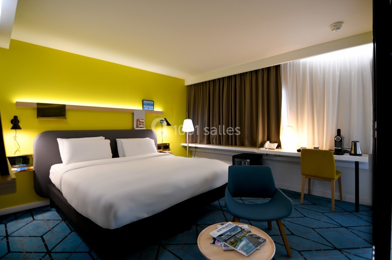 Chambre d'hôtel moderne avec lit double, bureau, fauteuil, éclairage chaleureux et murs jaunes.