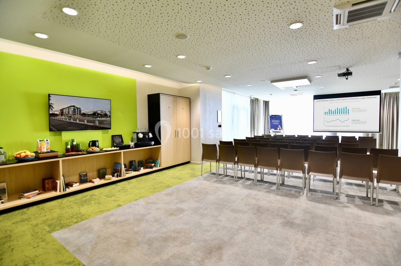 Salle de conférence lumineuse avec chaises alignées, écran de présentation, étagères et mur vert en arrière-plan.
