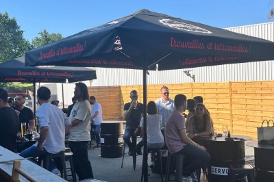 Terrasse couverte en bois avec tables hautes, tabourets noirs et deux personnes assises au fond près d'une barrière en bois.