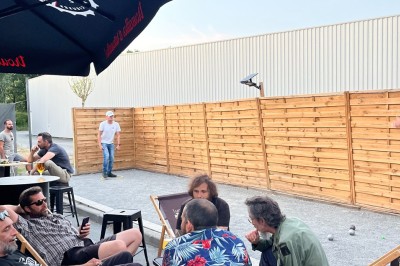 Terrasse couverte en bois avec tables hautes, tabourets noirs et deux personnes assises au fond près d'une barrière en bois.