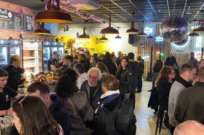 Terrasse couverte en bois avec tables hautes, tabourets noirs et deux personnes assises au fond près d'une barrière en bois.