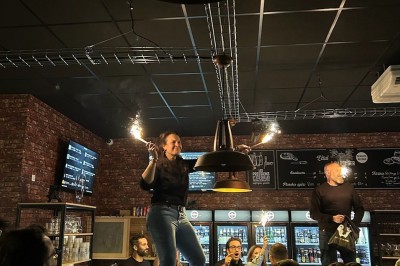 Terrasse couverte en bois avec tables hautes, tabourets noirs et deux personnes assises au fond près d'une barrière en bois.