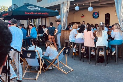Terrasse couverte en bois avec tables hautes, tabourets noirs et deux personnes assises au fond près d'une barrière en bois.