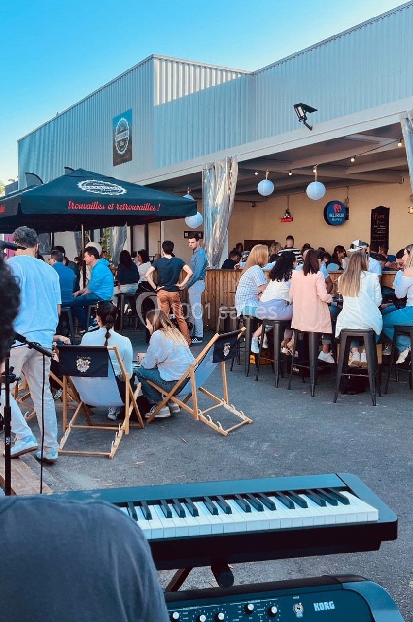 Groupe de personnes assises et debout dans une ambiance conviviale en extérieur près d'un bar, avec un clavier au premier…