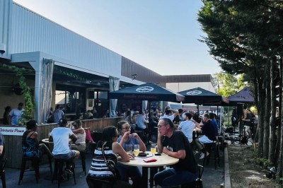 Terrasse couverte en bois avec tables hautes, tabourets noirs et deux personnes assises au fond près d'une barrière en bois.