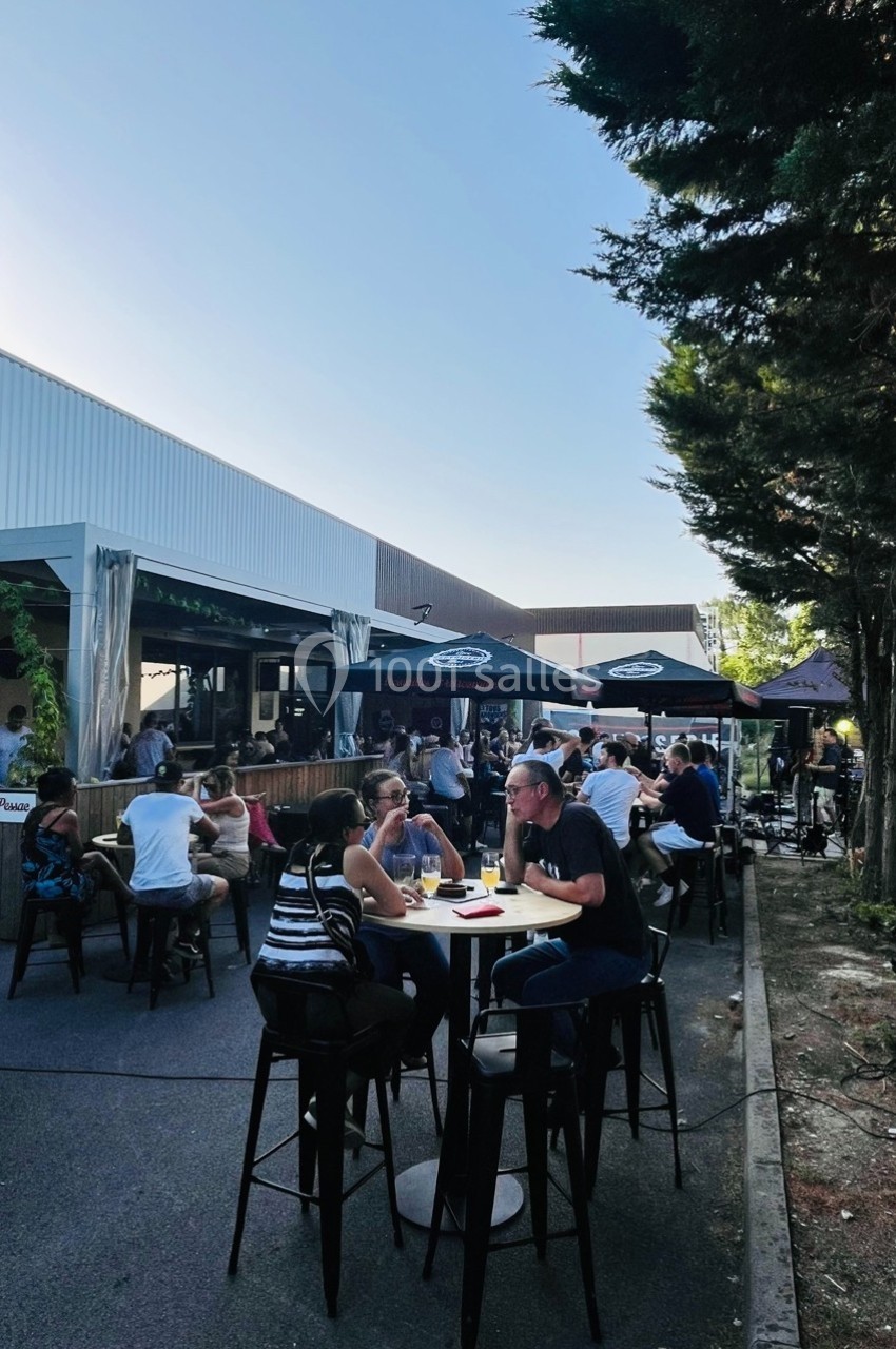 Personnes attablées en extérieur dans un espace de restauration avec parasols, arbres et ciel dégagé en arrière-plan.