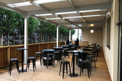 Terrasse couverte en bois avec tables hautes, tabourets noirs et deux personnes assises au fond près d'une barrière en bois.