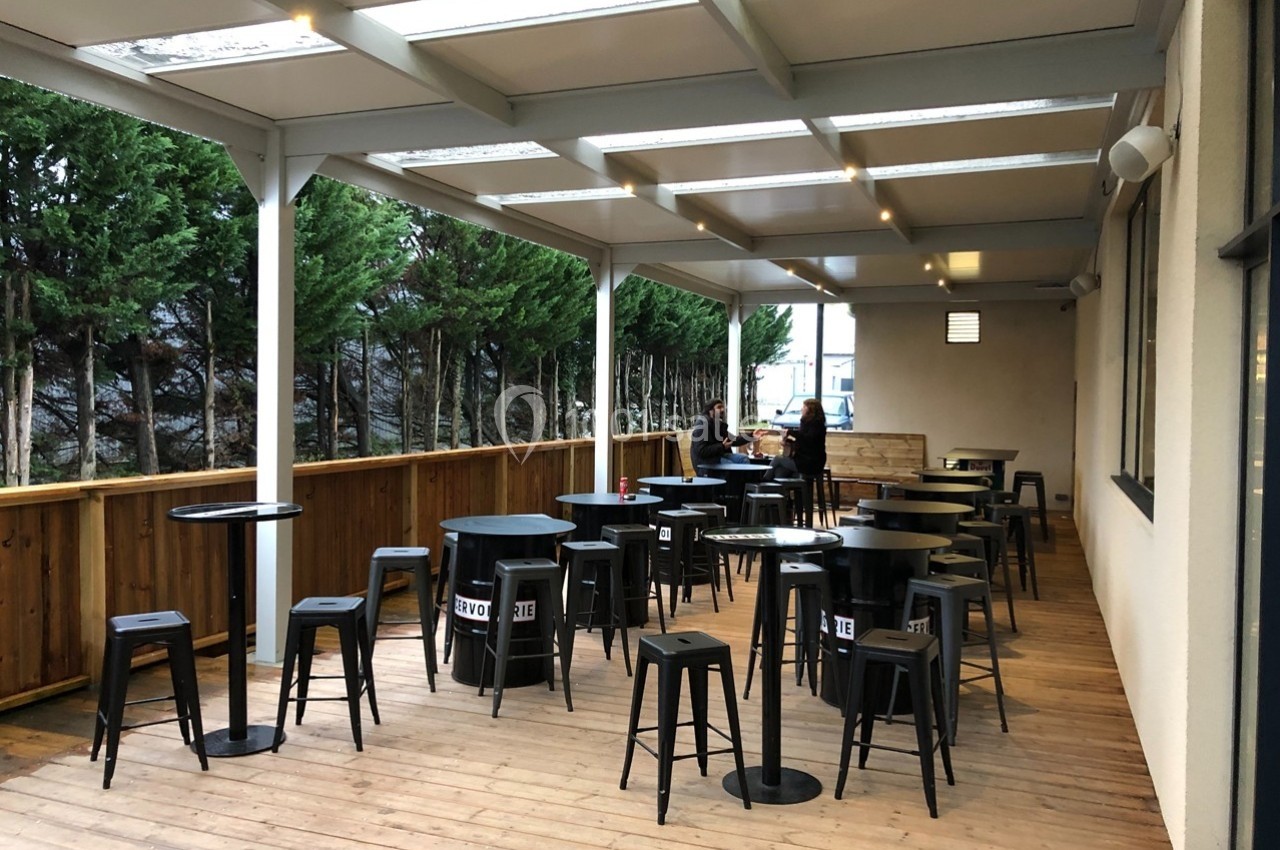 Terrasse couverte en bois avec tables hautes, tabourets noirs et deux personnes assises au fond près d'une barrière en bois.
