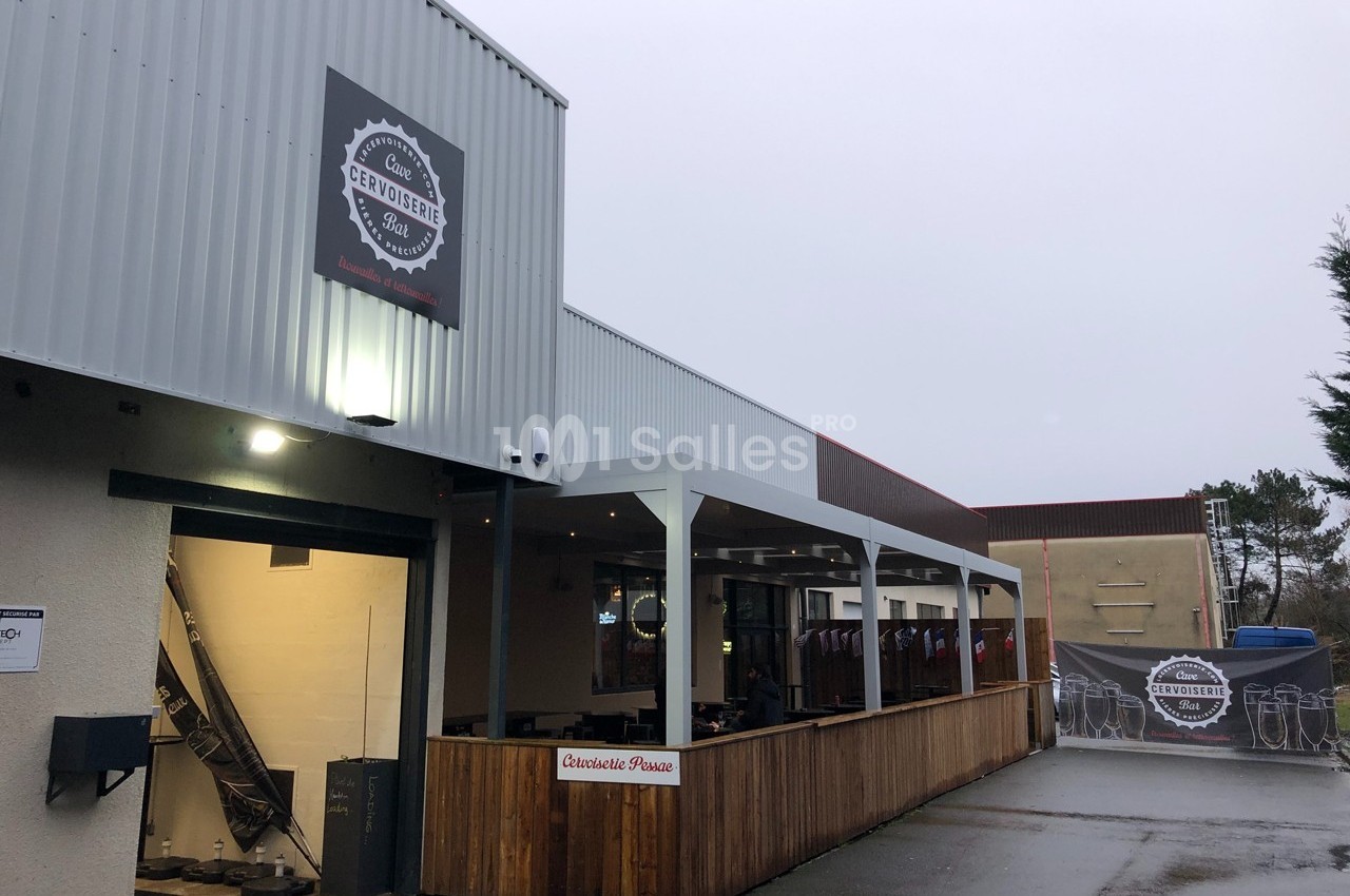 Façade d'une brasserie avec terrasse couverte en bois, éclairage extérieur et enseigne visible sous un ciel gris.