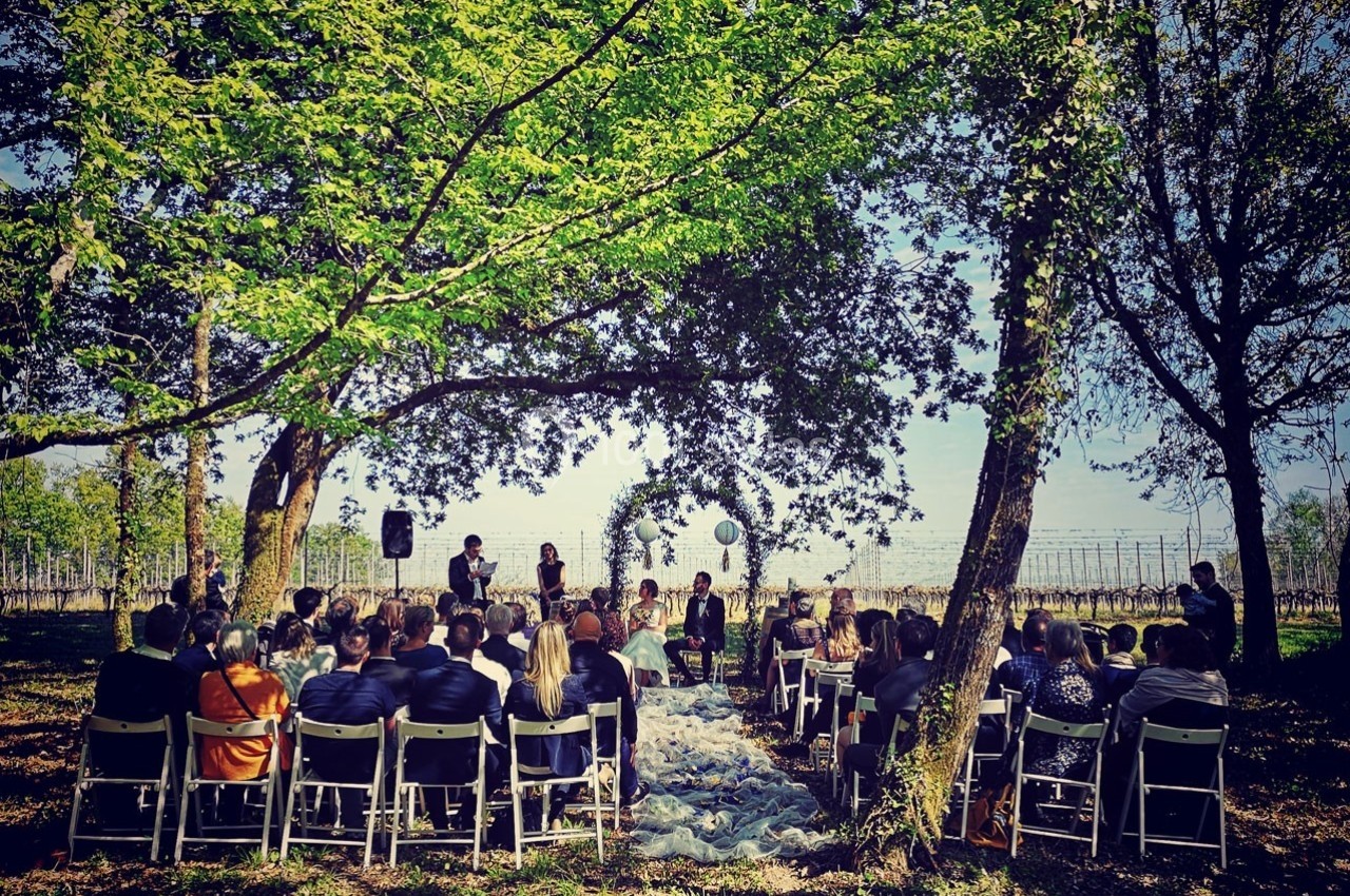 Cérémonie en plein air sous des arbres, avec des invités assis et un couple devant un arche décoré.