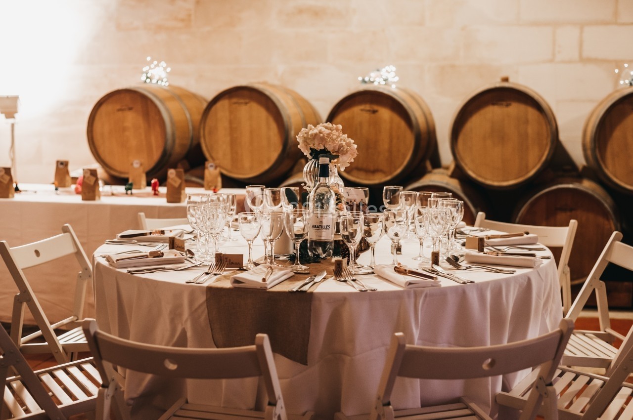 Table dressée avec vaisselle élégante, fleurs et bouteille, dans une salle avec des tonneaux en arrière-plan.